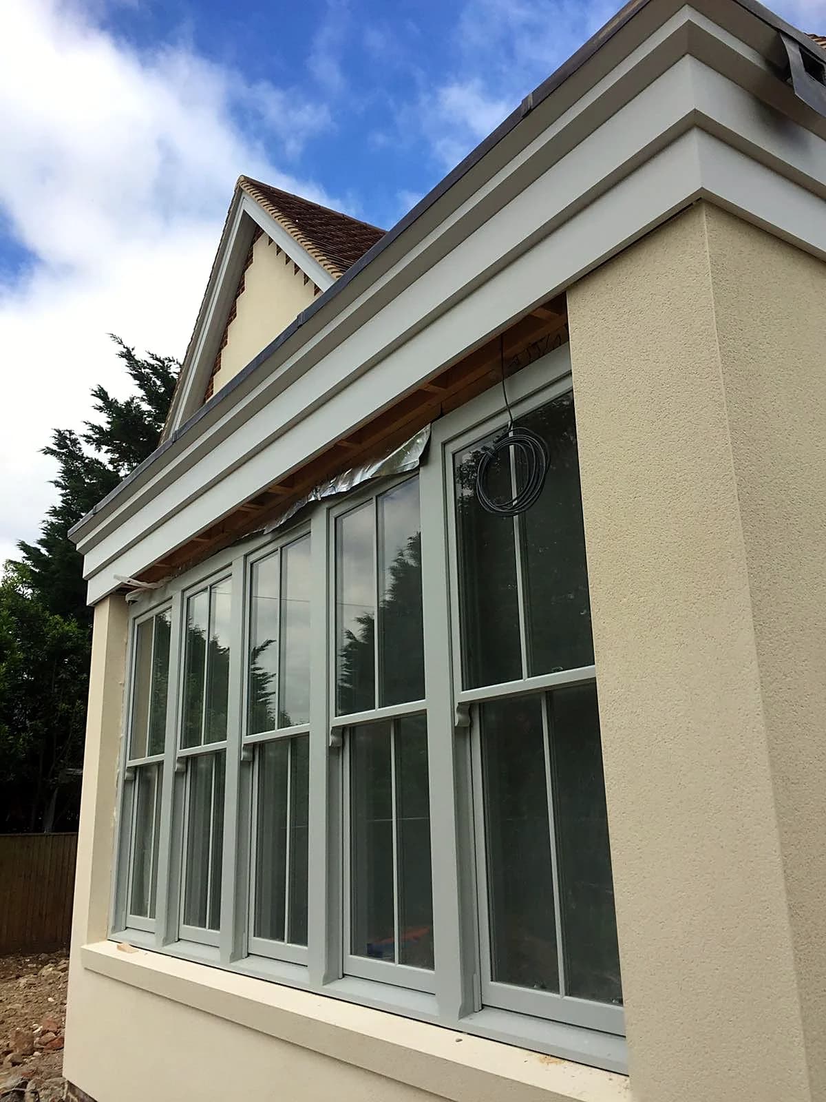 Corner of the new-build garden room with tall light grey timber sash windows against blue sky